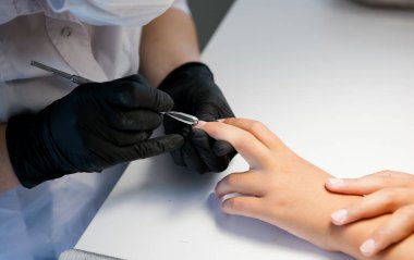 Siyah eldivenli bir usta bir kız için kerpetenle klasik bir manikür yapar. Beyaz ve sarı yüzüklü kadın elleri