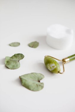 Yeşil yüz masajı silindiri, beyaz pamuk pedler, beyaz arka planda okaliptüs yaprakları.