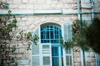 Eski bir taş binanın ağaç dalları tarafından çerçevelenmiş mavi pencere çerçeveleri ve panjurları
