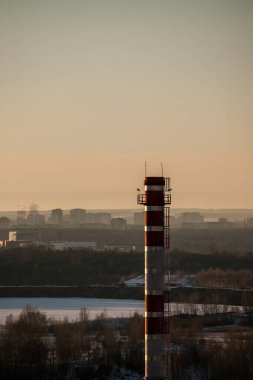 Parlak turuncu günbatımı binaları, sanayi bölgesini ve karla kaplı bir nehri aydınlatıyor.