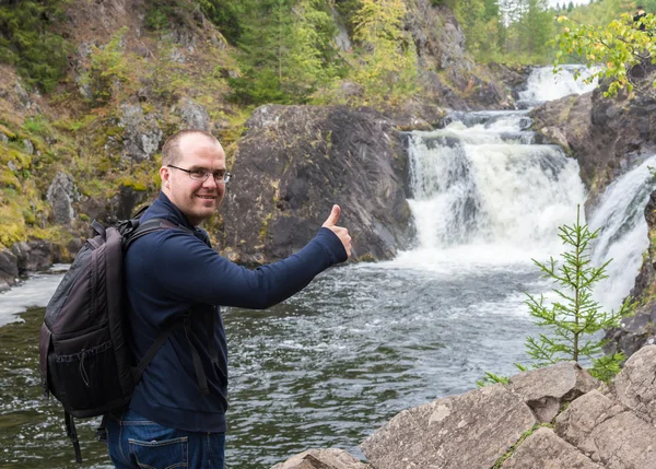 Homem com mochila dando polegar no fundo de uma cachoeira — Fotografia de Stock