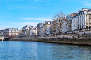 Paris 'teki sakin nehir manzarası. Sen nehri boyunca klasik binalar, zarif cepheler, mantarlı çatılar ve solda taş bir köprü. Açık mavi gökyüzü ve çıplak ağaçlar barışçıl bir ilkbahar atmosferi yaratır.