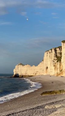 Sıcak günbatımı ışığında, insanlar çakıl taşı plajı boyunca yürüyorlar ve ünlü Etretat kayalıklarının altında dalgalar kıyıya ulaşıyor. Manzaralı Normandiya kıyıları ikonik doğal işaretli ve akşam seyahati atmosferli.
