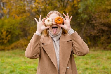 Kız yüzünün bir kısmını turuncu ve beyaz balkabaklarıyla kaplıyor. Hasat konsepti. Uzayı kopyala Gülümseyen kadın şakacı davranarak gözlerini balkabağıyla kapatıyor.