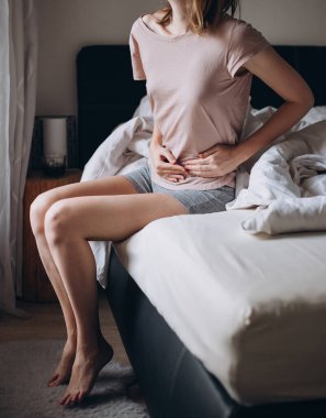 Güçlü karın ağrısı çeken genç bir kadın, sıradan bir ev çekimi. Yatakta oturan kadın.