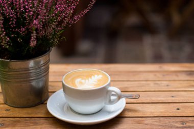 Bir fincan kapuçino, ahşap masa üzerinde latte sanatı, beyaz seramik bardak, yazı için yer. Kafede ya da evde masası olan teras.