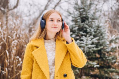 Kulaklıkla müzik dinleyen mutlu genç bir kadın. 2021 yılının modaya uygun rengi. Aydınlatıcı sarı. Göz kamaştırıcı kış manzaraları arasında seyahat ederken.