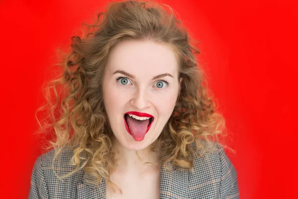 Portrait of joyful pretty woman over red background, emotional curly girl showing tongue at the camera. Emotion facial expression. — Stock Photo, Image