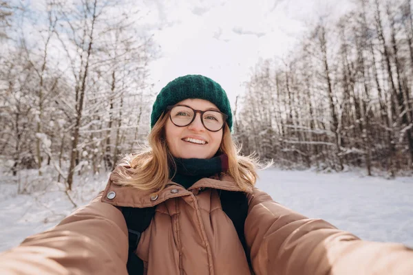 녹색 겨울에 뜨개질 모자를 쓴 행복 한 아가씨는 겨울 숲에서 셀카를 먹는다. 카메라를 보고 웃고 있죠. 여행과 활동적 인 삶의 개념. 옥외에서 — 스톡 사진