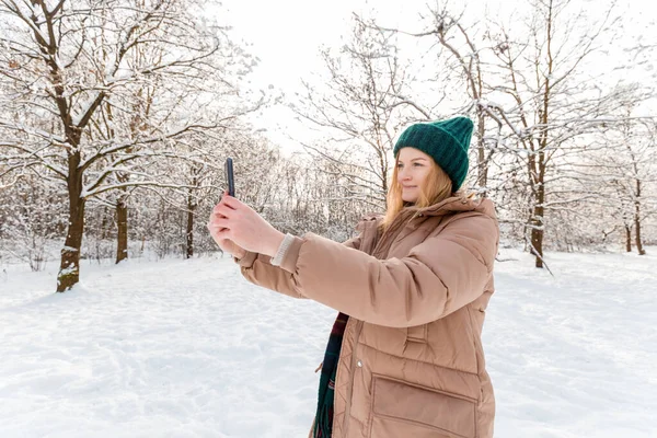 Mladá krásná šťastná dívka v zimě pletený klobouk se selfie v zimním lese. Žena pomocí aplikace chytrý telefon přes přírodu pozadí — Stock fotografie