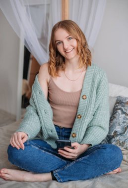 Genç, güzel, mutlu bir kız rahat bir yatağa mesaj yazıyor. Akıllı telefon uygulaması kullanan bir kadın. Çevrimiçi iletişim kavramı.