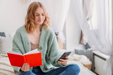 Portrait of a pretty young blond girl hiding behind an open orange book and phone. Happy Woman studying or working on cozy bed. Home schooling concept