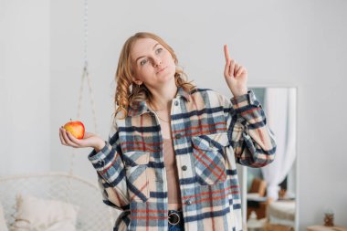 Genç, güzel, sarışın bir kadın elinde kırmızı elmayla arka planda olanları düşünüyor. Sağlıklı beslenme, vejetaryenlik.