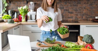 Beyaz tişörtlü kadın ev mutfağında hareketli salata pişiriyor. Sağlıklı yemek pişirme süreci. Menü, yemek tarifi kitabı afişi. Kız vegan salatası yaparken video izlemek için dizüstü bilgisayar kullanıyor