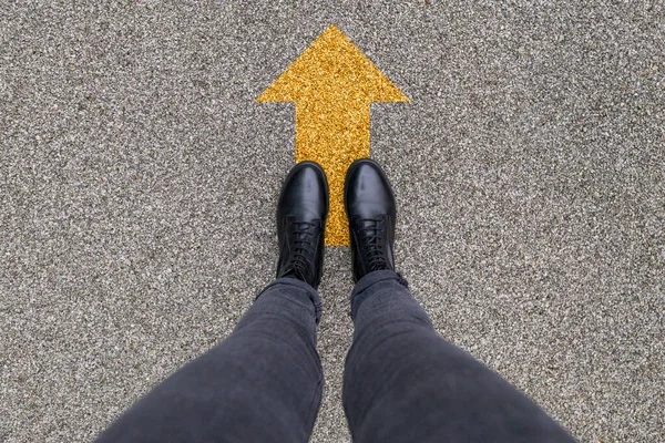 Black shoes standing on the asphalt concrete floor with yellow direction arrow symbol. Moving forward, new start and success.. Feet shoes walking in outdoor. Youth Selphie Modern hipster