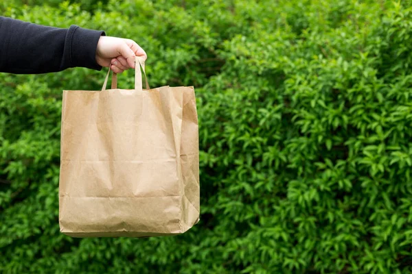 Close up de homem mão com saco de papel para levar comida no fundo verde natureza. Entrega em qualquer clima 24 horas por dia para o cliente. — Fotografia de Stock