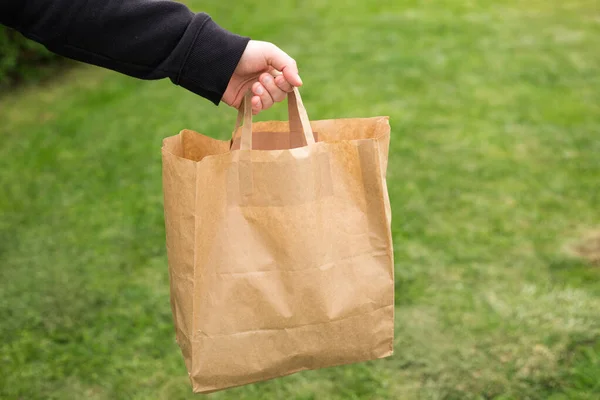 Close up de mão de homem com saco de papel ecológico para levar comida no fundo verde natureza. Entrega em qualquer clima 24 horas por dia para o cliente. — Fotografia de Stock