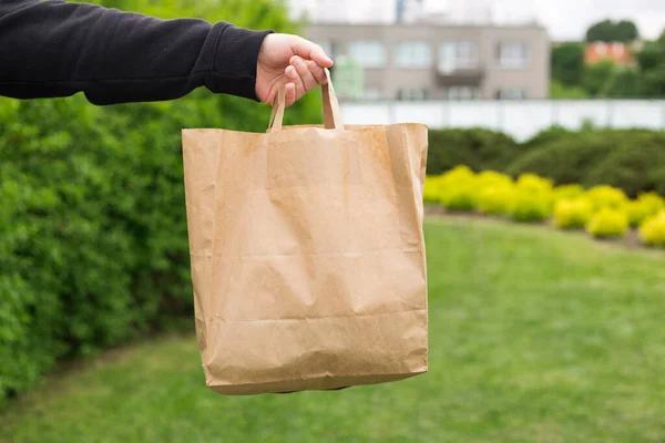 Close up de mão de homem com saco de papel ecológico para levar comida no fundo verde natureza. Entrega em qualquer clima 24 horas por dia para o cliente. — Fotografia de Stock