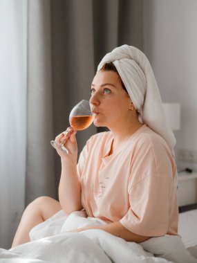 Duş aldıktan sonra yatak odasında bir bardak şarapla güzel, mutlu bir genç kadın. Karantina süresince evde tecrit altında kalmak. Doğum gününü yatakta yalnız kutlama kavramı