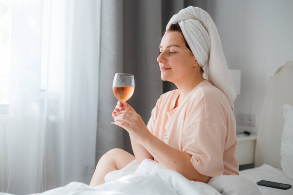 Красивая счастливая молодая женщина после душа с бокалом вина в спальне. Оставаться дома в изоляции во время карантинной изоляции. Концепция празднования дня рождения в постели в одиночку