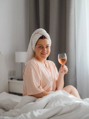 Duş aldıktan sonra yatak odasında bir bardak şarapla güzel, mutlu bir genç kadın. Karantina süresince evde tecrit altında kalmak. Doğum gününü yatakta yalnız kutlama kavramı