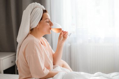 Duş aldıktan sonra yatak odasında bir bardak şarapla güzel, mutlu bir genç kadın. Karantina süresince evde tecrit altında kalmak. Doğum gününü yatakta yalnız kutlama kavramı