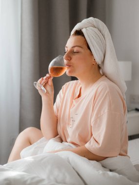 Duş aldıktan sonra yatak odasında bir bardak şarapla güzel, mutlu bir genç kadın. Karantina süresince evde tecrit altında kalmak. Doğum gününü yatakta yalnız kutlama kavramı