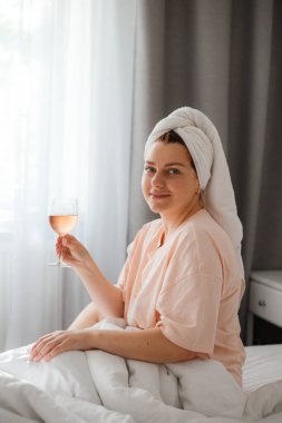 Duş aldıktan sonra yatak odasında bir bardak şarapla güzel, mutlu bir genç kadın. Karantina süresince evde tecrit altında kalmak. Doğum gününü yatakta yalnız kutlama kavramı
