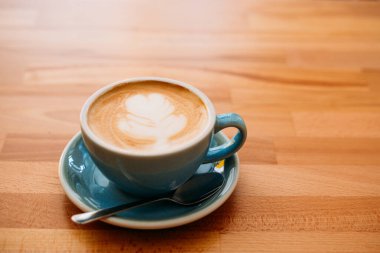 Latte sanatıyla birlikte bir bardak kapuçino. Kafedeki ahşap masada mavi bardakta kahve.