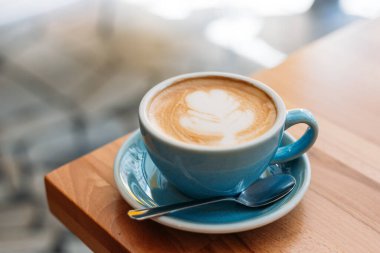 Latte sanatıyla birlikte bir bardak kapuçino. Kafedeki ahşap masada mavi bardakta kahve.