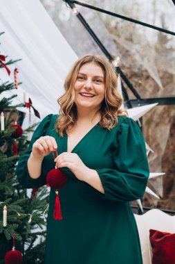 Yeşil elbiseli genç bir kadın evde Noel ağacı süslüyor ve gülümsüyor. Kış ve tatiller. Kutlama bayrağı.