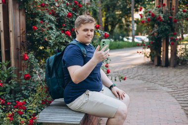 Sarışın, Avrupalı 30 'lu yaşlarda bir adam parkta müzik dinleyip eğleniyor. Yakışıklı bir erkek bankta oturuyor ve Bluetooth kulaklıkla müzik dinliyor. Yaşam tarzı, teknoloji, insan kavramı.