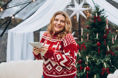 Gülümseyen kadın dijital tablete bakıyor ve süslü Noel ağacının yanında duruyor. Kış ve tatiller. Kutlama afişi, büyük indirim.
