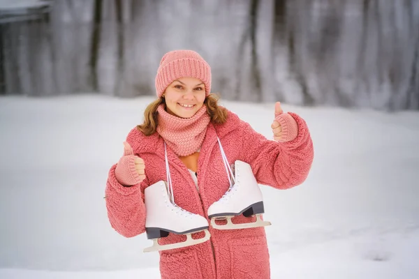 ピンクのエコファーのコートを着た若い女性は冬の日に肩に白いスケートを持っています 冬は屋外でアイススケートをします 健康的なライフスタイルのコンセプト ロイヤリティフリーのストック写真