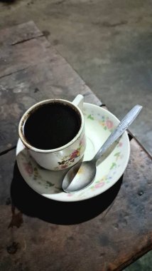 Beyaz çiçekli bir fincanda koyu siyah kahve, desenli, aşınmış ahşap bir masada kaşık, sıcacık bir sabahı çağrıştırıyor..