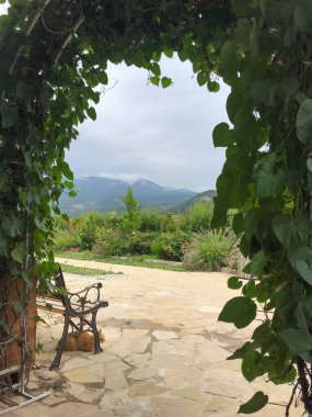Bereketli sarmaşıklarla kaplı sakin bir bahçe kemeri, yumuşak, bulutlu bir gökyüzünün altındaki uzak dağlara bakan, işlenmiş demir bir bankla, renkli çiçeklerle dolu bir terası çerçeveler..