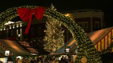 Riga, Letonya - 4 Aralık 2025: Dome Meydanı 'ndaki Noel pazarı girişi aydınlık bir kemer ve büyük kırmızı bir fiyonk, şenlik tezgahları ve akşam gökyüzünün altında parlak ışıklandırılmış bir Noel ağacı. 