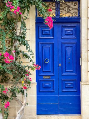 Valletta, Malta 03-oct-2024. Yeşil sarmaşıklar ve canlı pembe begonviller tarafından çerçevelenmiş, açık taştan duvarda duran çarpıcı mavi kapı. Süslü demircilik Akdeniz tarzı girişe cazibe katıyor.
