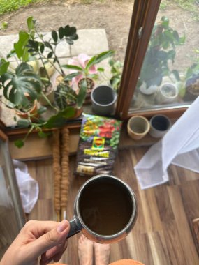 Wroclaw, Polonya - 11 Ekim 2025: Bir el, çeşitli saksı bitkileri ve bahçe malzemeleriyle dolu kapalı bir bahçede bir fincan çay tutuyor