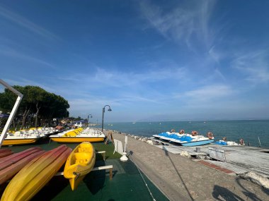 Sirmione, İtalya - 10 Ekim 2025: Kıyı boyunca renkli kanolar ve pedallı botlar düzenlenirken, insanlar deniz kıyısındaki güneşli günün tadını çıkarıyorlar