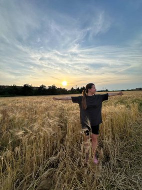 Büyük beden gömlekli kadın figürü altın buğdayların arasında duruyor, kolları açık batan güneşe doğru, sükunet ve doğal güzelliği yakalıyor.