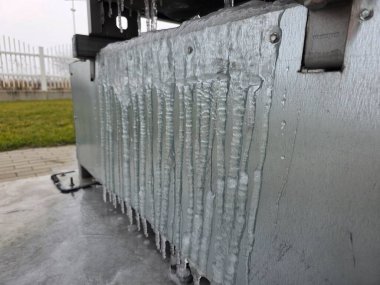 Buz oluşumları metalik bir yüzeyde serpintiler ve dokularla mevcuttur, soğuk hava koşullarında çimenli bir alanla zıt düşer ve arka planda beyaz bir çit bulunur.