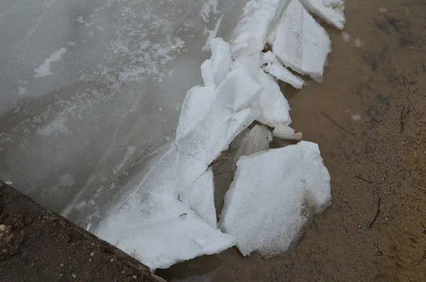 Kırık buz örtüleri kumlu nehir kıyısında dinleniyor. Su birikintileri ve eriyen buzlar görülebiliyor. Kış manzarası yaratıyor.