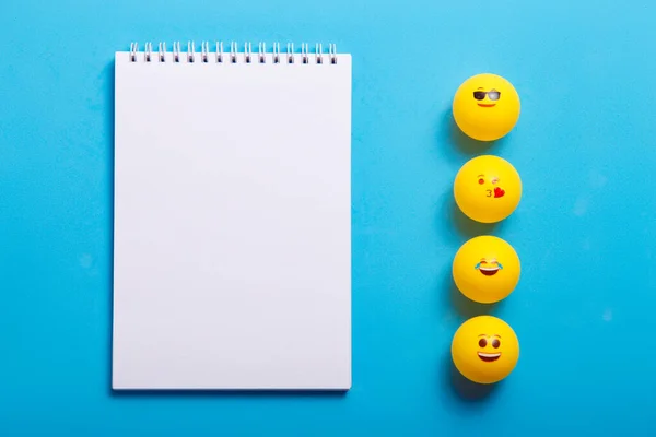 Mavi arka planda farklı emojileri olan birkaç sarı top ve not defteri.