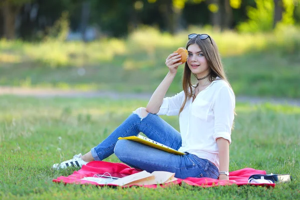 Giovane ragazza seduta sull'erba nel parco e lavora in un computer portatile mangiare fast food — Foto Stock