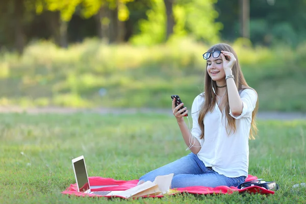 Jeune fille assise dans le parc, souriant et travaillant à son ordinateur portable, écoutant de la musique — Photo