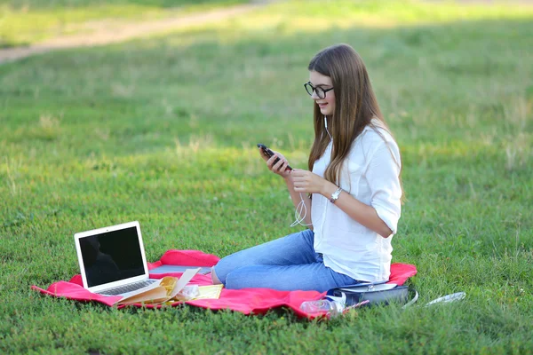 Jeune fille assise dans le parc, souriant et travaillant à son ordinateur portable, écoutant de la musique — Photo