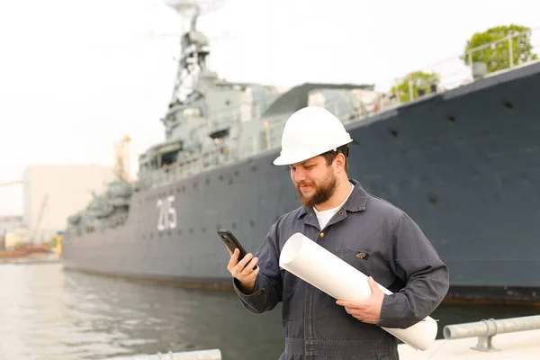 Marine engineer talking by VHF walkie talkie, holding papers near vessel in background.