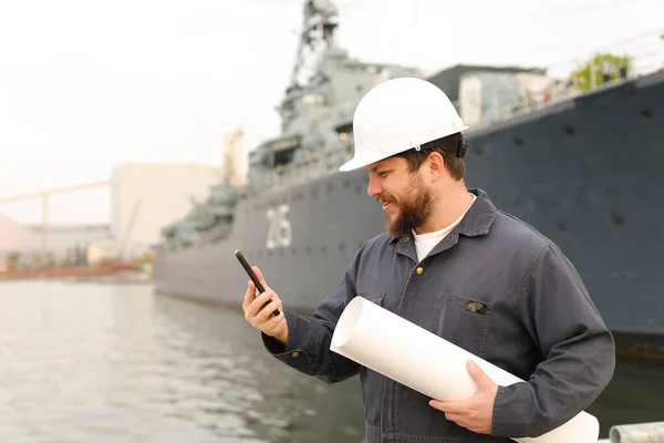 Marine DDE, designated duty engineer, talking by VHF walkie talkie, holding papers near vessel in background.