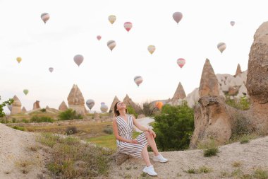 Taşın üzerinde oturan gülümseyen kız, arka planda sıcak hava, Kapadokya.
